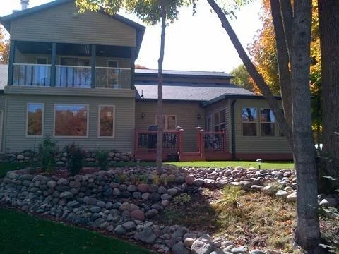 Backyard summer view featuring the creekside living room area on left, with upper level covered balcony above, and the 3-season porch on the right, with a private deck in between for grilling or just enjoying the peace and quiet here!