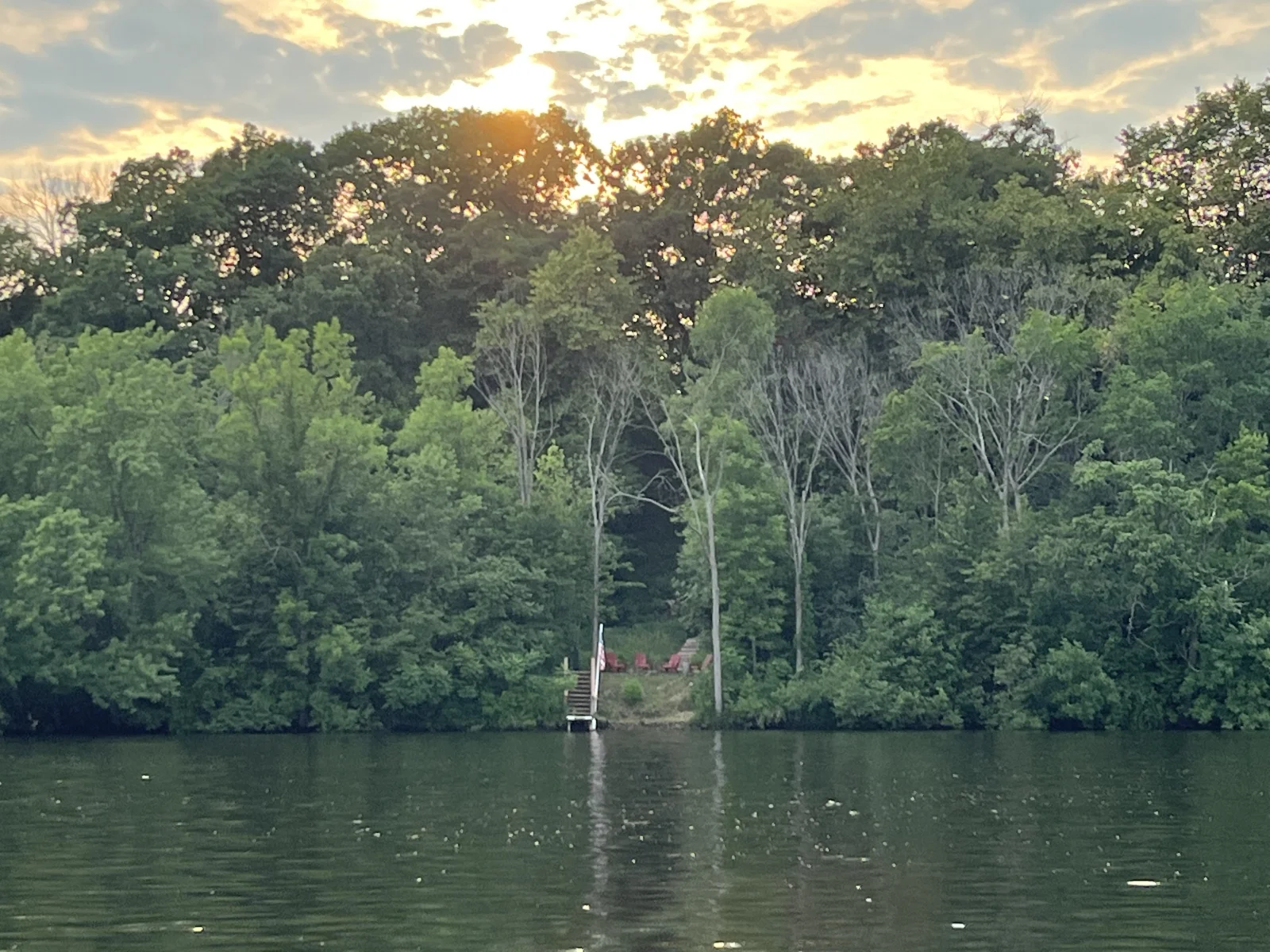 A view of the property from across the river. The dock and red lawn chairs are visible on the shore beyond.