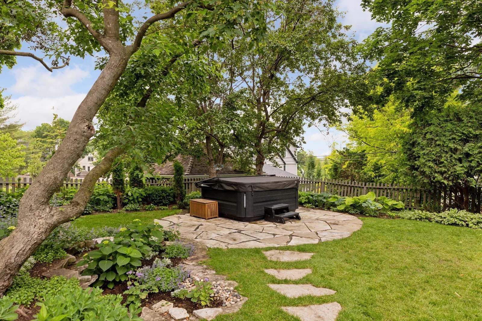 Stone pavers lead to a stone paver patio and hot tub surrounded by manicured gardens and mature hedges. Seasonal lake views.