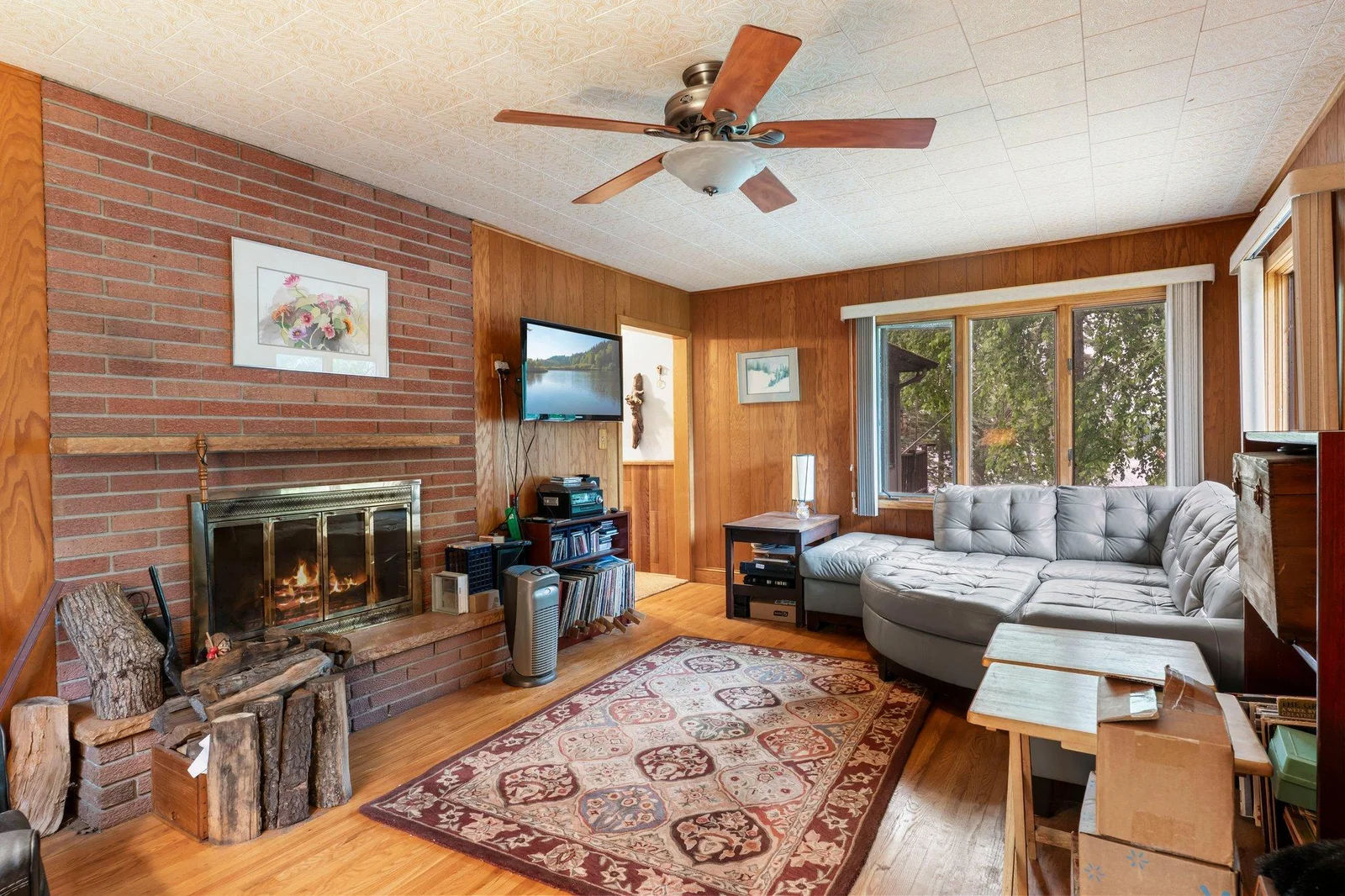Step into the inviting main living room, where rich wood flooring, classic wood-paneled walls, and a cozy wood-burning fireplace with full brick surround create a warm and timeless atmosphere. A large closet offers convenient storage.