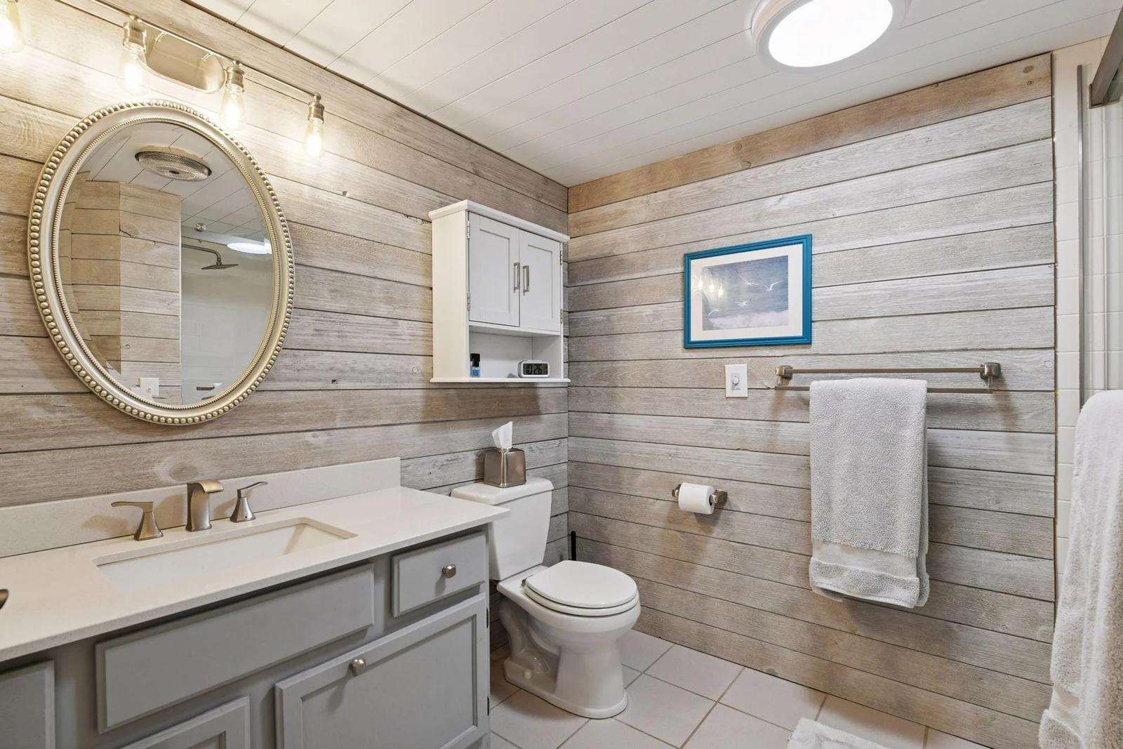 This bathroom exudes a perfect lake house vibe, featuring shiplap walls/ceiling, tiled floors, a round portal mirror, and a stunning walk-in shower with tile surround. A fun and refreshing space to unwind and pamper yourself after a day outdoors.