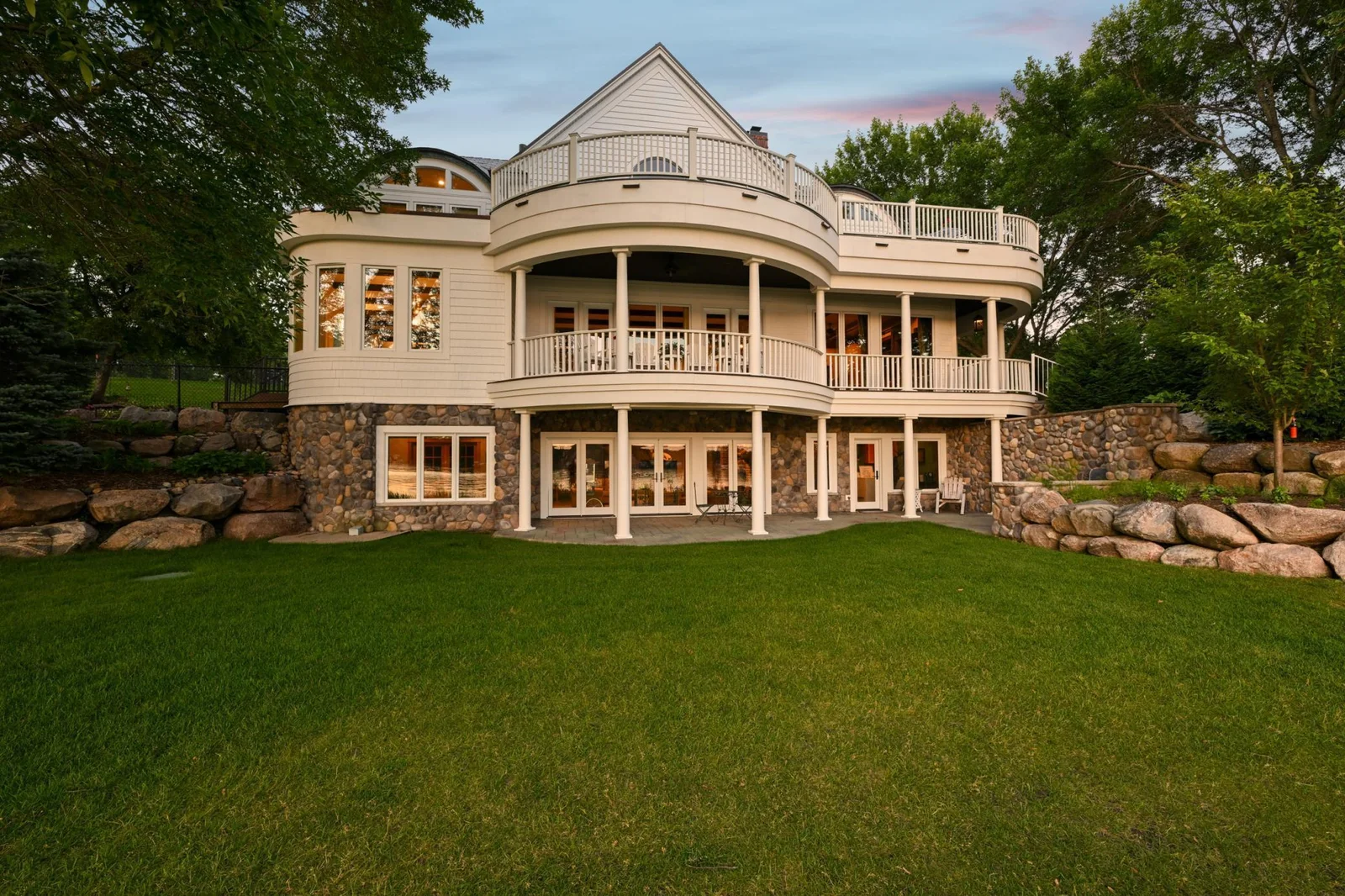 This lakeside view highlights the home’s elegant architecture with three levels of covered outdoor living, natural stonework, and beautifully landscaped grounds just steps from the water’s edge.