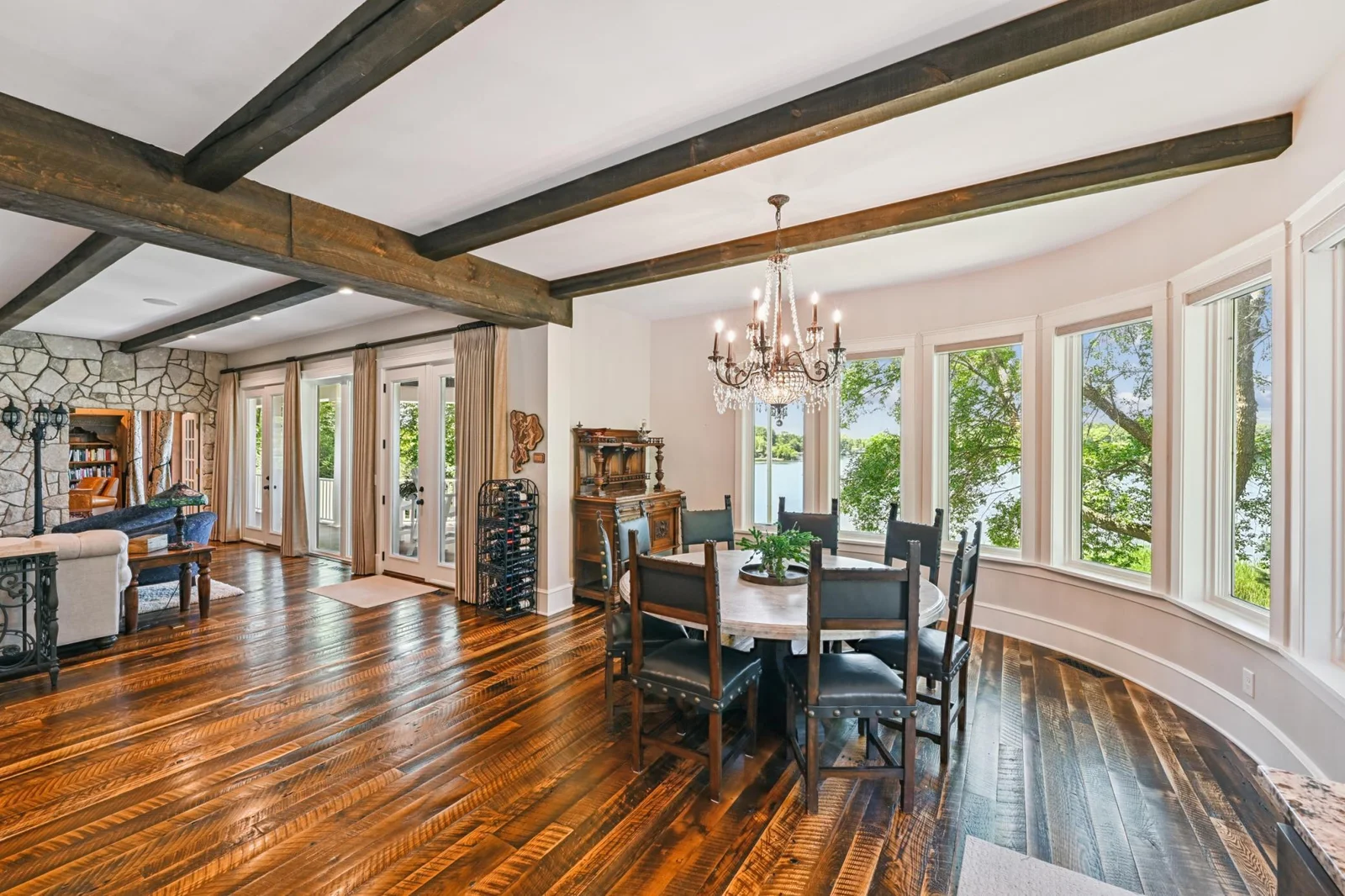 Elegant dining area framed by a curved wall of lakeside windows with panoramic water views, rich hardwood floors, exposed ceiling beams, and a sparkling chandelier, perfect for memorable meals by the lake.