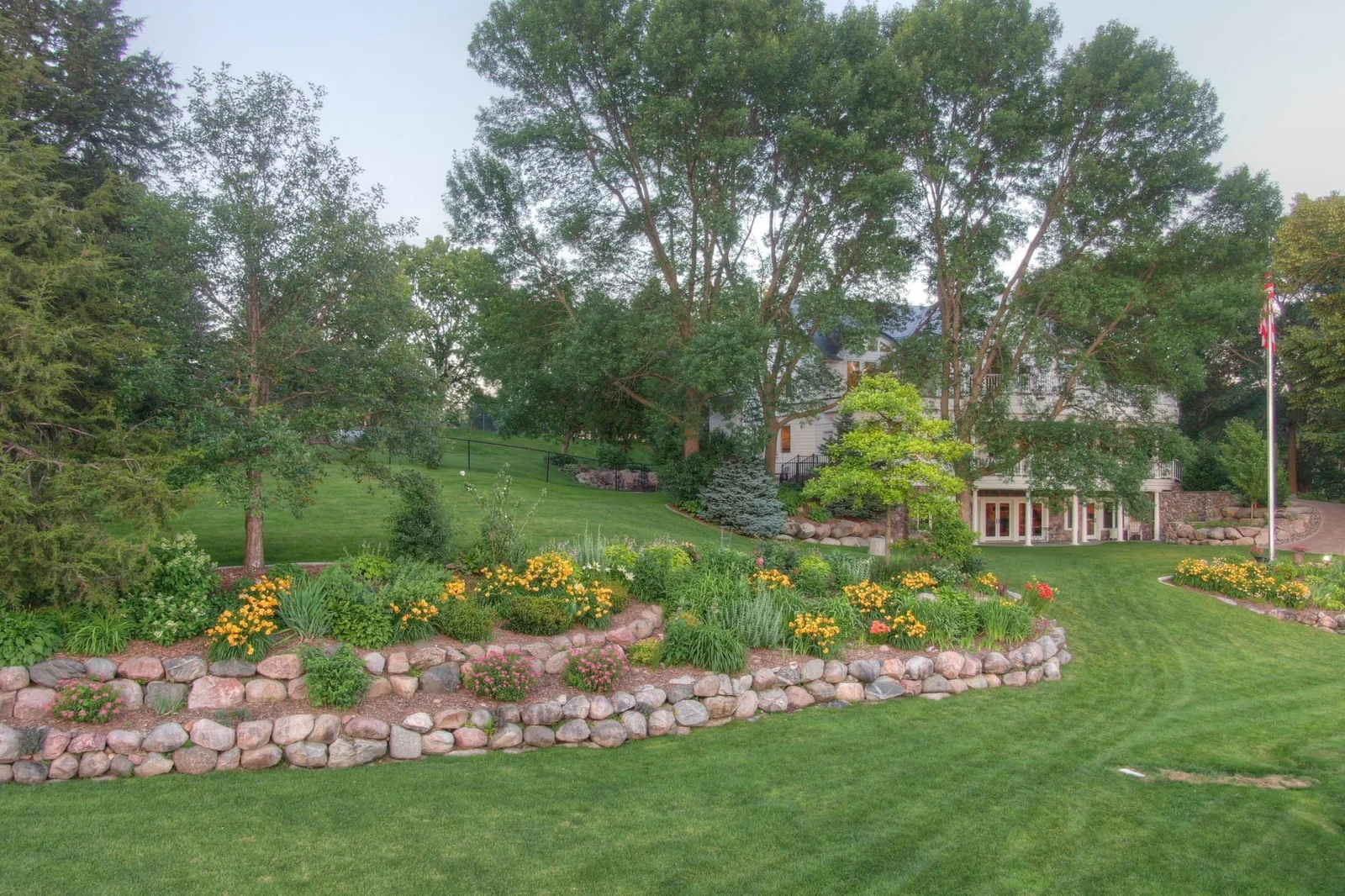 Professionally landscaped tiered garden featuring vibrant perennials, natural stone retaining walls, and mature trees that frame the estate with beauty and privacy. All sides of the property other than the shoreline are fenced in.