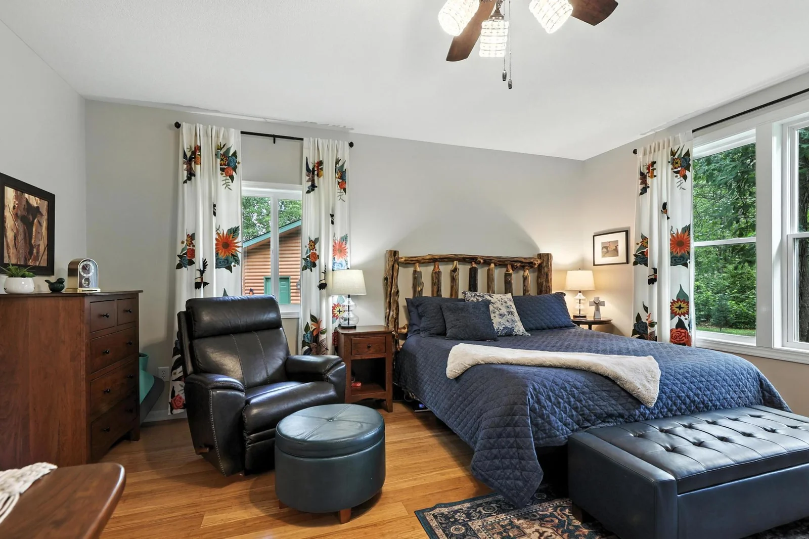 This spacious primary bedroom offers a peaceful woodland view through oversized windows and features rich wood flooring, soft neutral tones, and custom floral drapes that complement the rustic wood headboard.