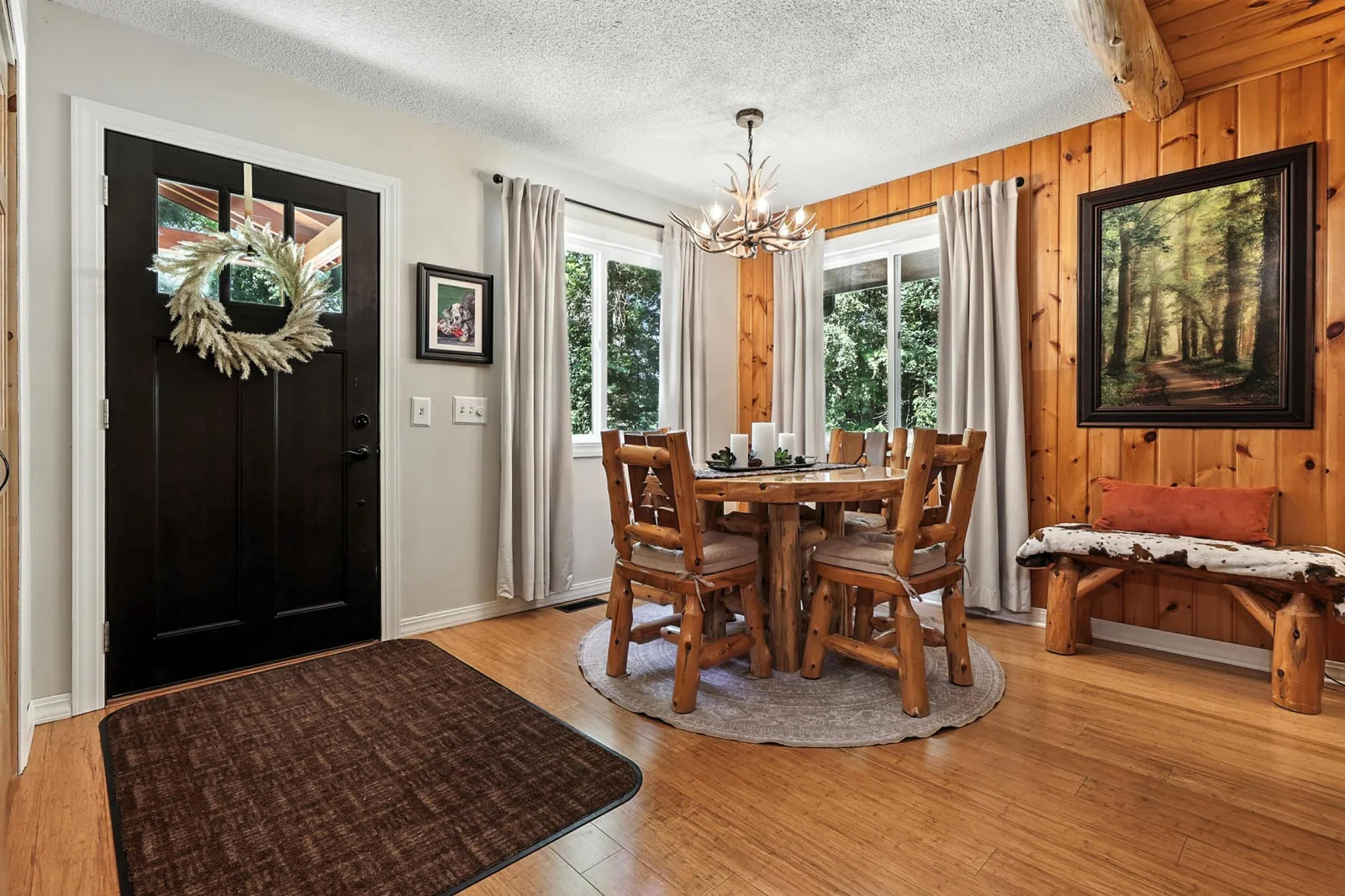 Warm and welcoming entry with natural light streaming through dual windows. The adjacent dining space features wood paneling, a rustic antler chandelier, and charming log furniture for that cozy cabin feel.