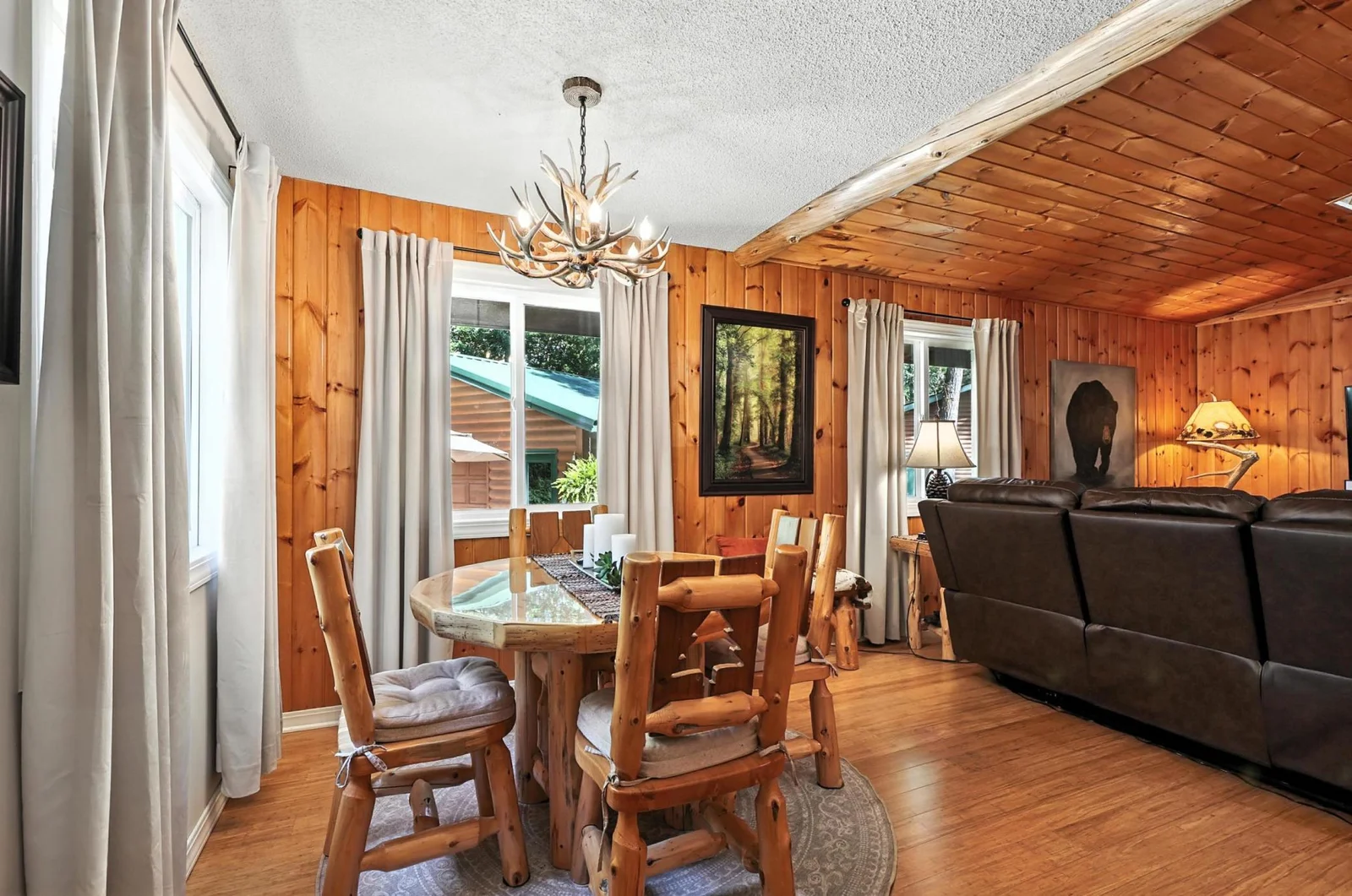 Surrounded by windows, this bright dining area blends into the living space with wood-paneled walls and ceiling, neutral curtains, and a lodge-style ambiance ideal for entertaining.