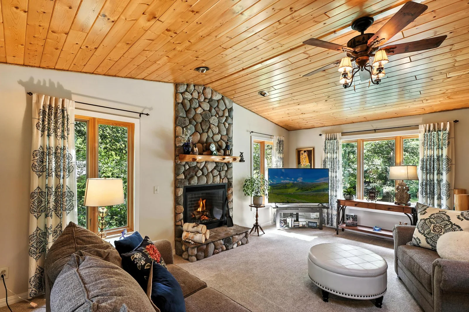 Warm and welcoming space featuring vaulted wood ceiling, large windows, and a stunning river rock fireplace.