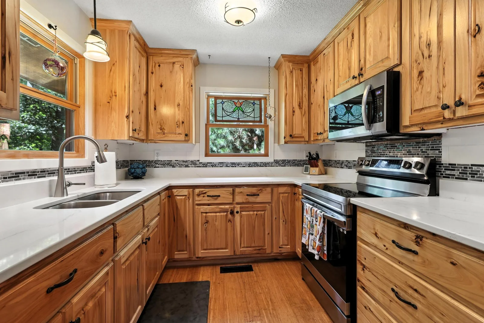 Beautifully updated kitchen with knotty wood cabinets, white quartz countertops, sleek stainless-steel appliances, and a mosaic tile backsplash. A decorative stained-glass window above the sink adds charm.