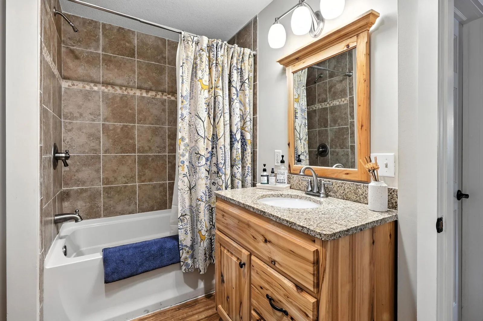 This full bathroom boasts a rustic charm with a natural wood vanity, granite countertop, and tiled shower surround, enhanced by warm lighting and playful patterned shower curtain.